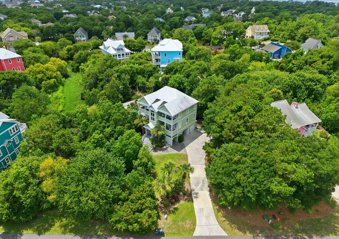 Aerial view of a charming coastal neighborhood featuring a green cottage surrounded by lush summer foliage, with glimpses of waterfront beyond.