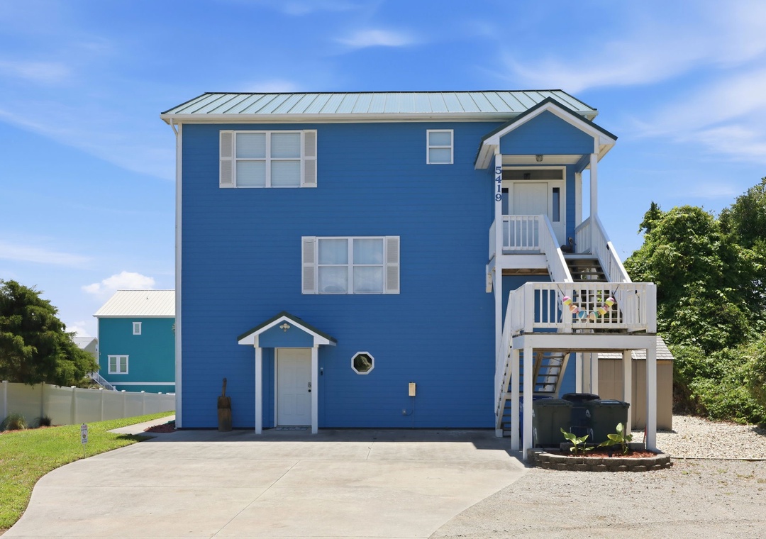 This charming blue coastal home features elevated architecture with private balconies, convenient ground-level parking, and a peaceful residential setting surrounded by natural greenery.