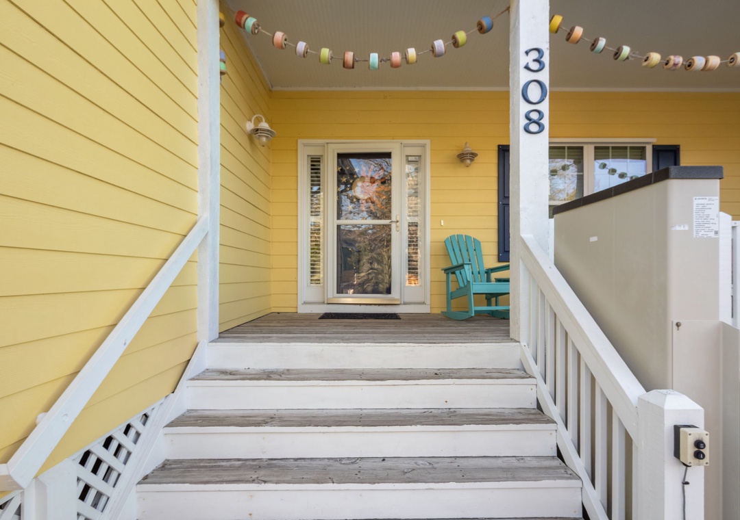 Charming yellow cottage entrance welcomes you with cheerful bunting and a cozy turquoise chair on the front porch.