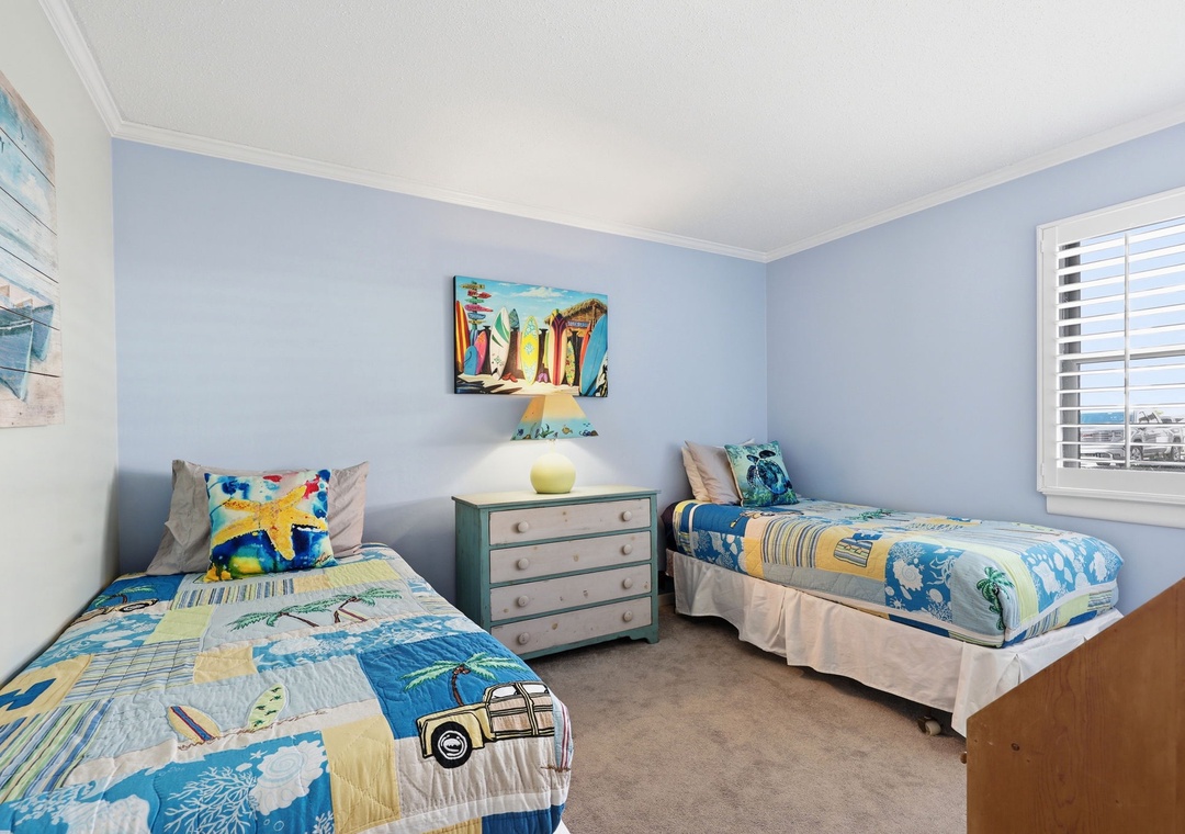 Your beachside bedroom features two twin beds with coastal-inspired bedding, soft blue walls, and cheerful surfboard artwork that sets a playful, relaxing vacation tone.