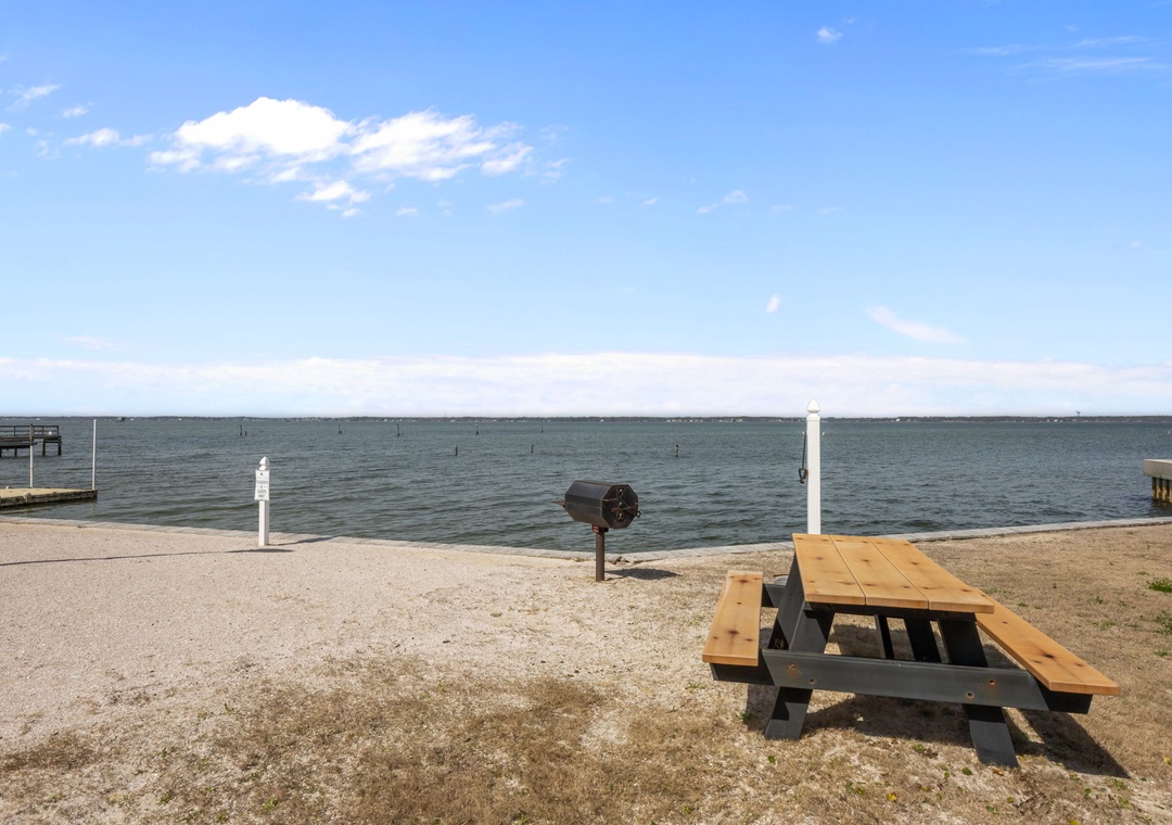 Waterfront picnic area with table and grill overlooking calm waters under blue skies.
