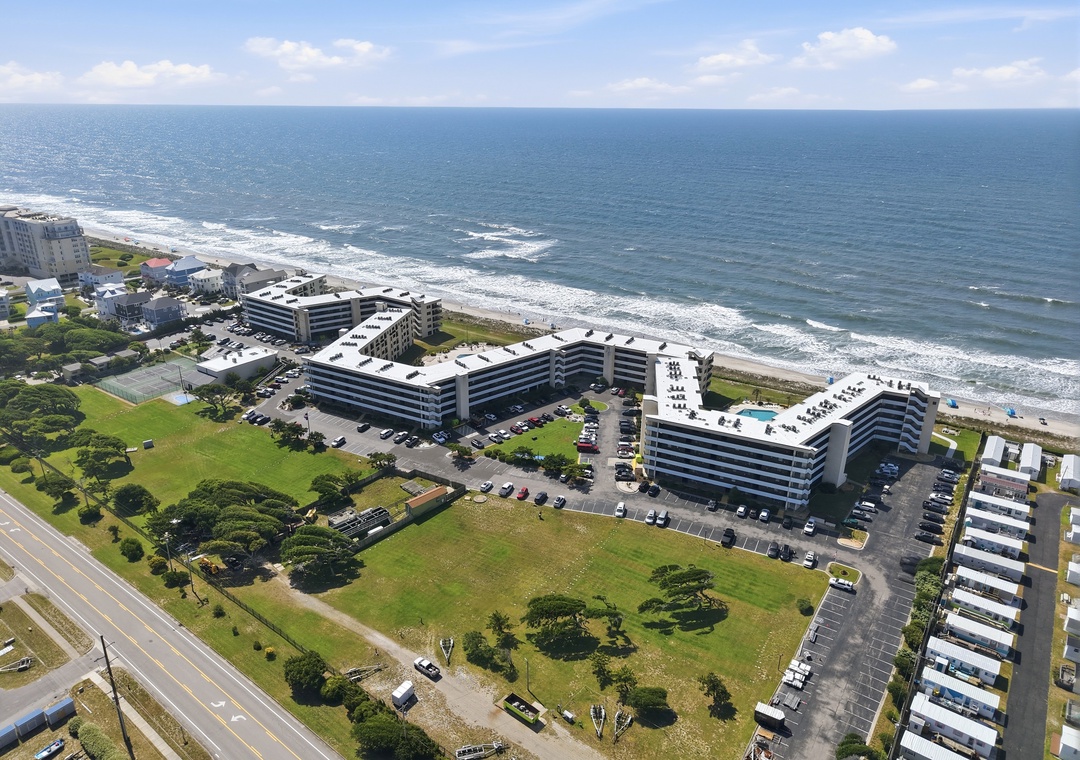 Aerial view of beachfront property with ocean access, green lawns, and modern multi-story buildings situated directly along the coast with rolling waves and sandy shoreline.