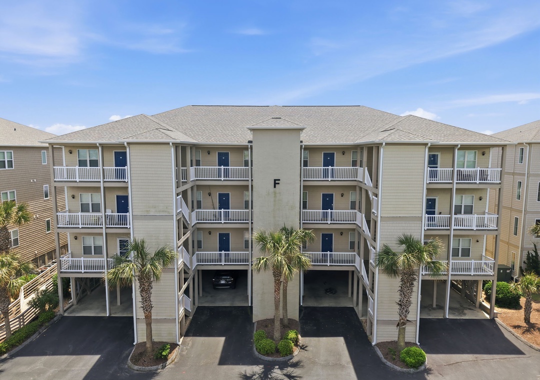 Multi-story beachside building featuring elevated parking and private balconies beneath swaying palms under a clear blue sky.