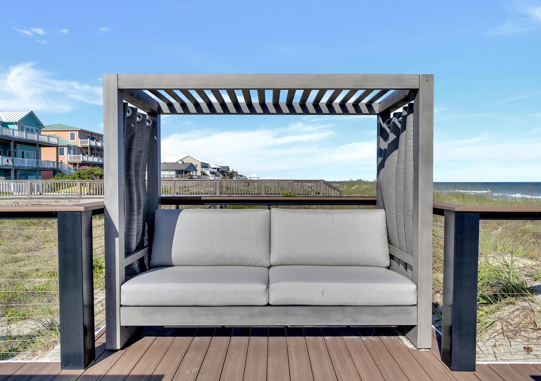 Oceanfront daybed with pergola shade on a private deck overlooking the beach and rolling waves, with direct beach access steps away.