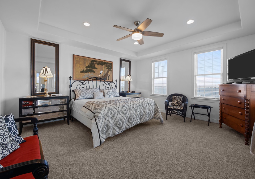 Relax in your elegant bedroom featuring a plush bed, ceiling fan, flat-screen TV, and natural light from two windows.