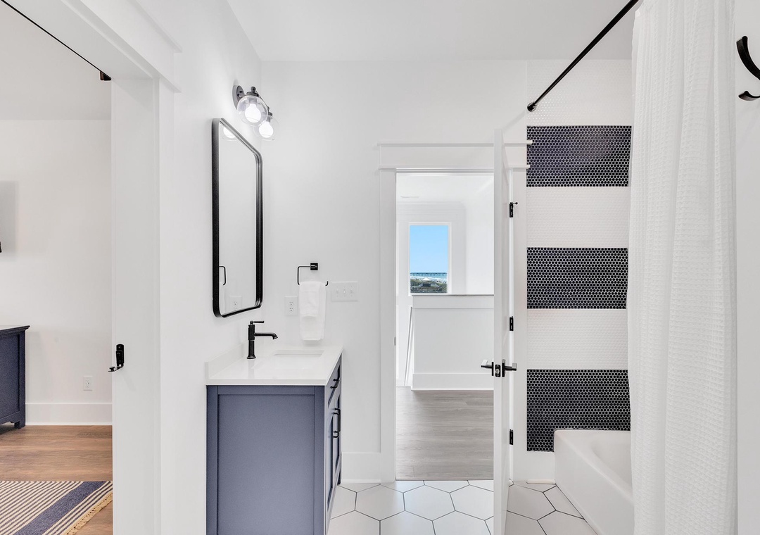 Your private bathroom features modern fixtures, sleek hexagon tile flooring, and a shower with striking black penny tile accents. Natural light flows through the adjacent room, adding warmth to the space.