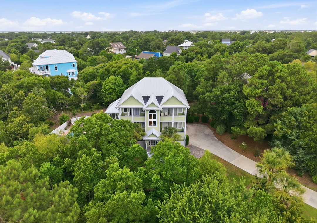 Aerial view showcases a spacious three-story coastal home with a metal roof, nestled among lush oak trees in a quiet residential neighborhood, with convenient parking and nearby community amenities.