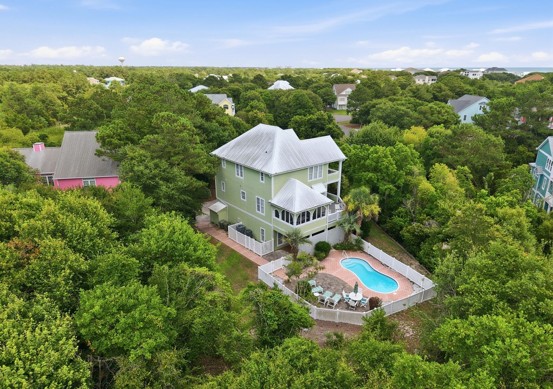 Aerial view of a spacious green vacation home nestled among coastal trees, featuring a private pool surrounded by lounge chairs, white fencing, and tiered decking in a quiet residential neighborhood.