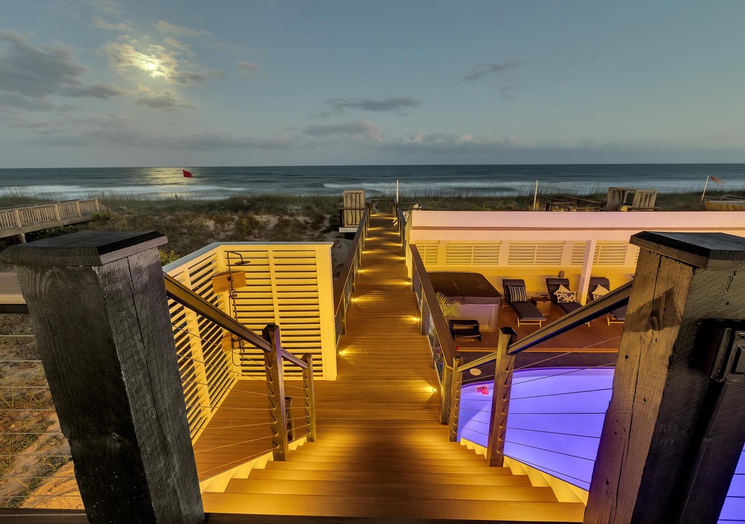 A beachfront multilevel deck with ambient lighting leads to the sandy shore and ocean. This elevated outdoor space features lounging areas and direct beach access for coastal relaxation.