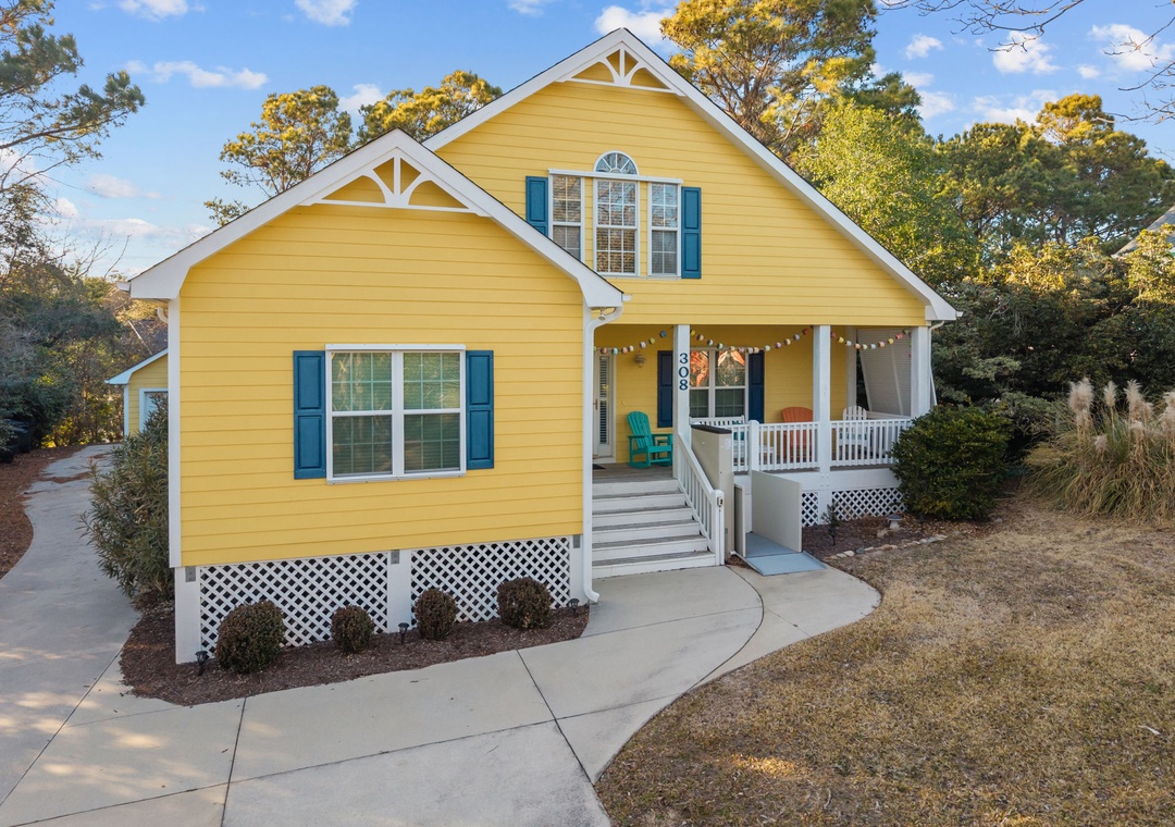 Charming yellow cottage with cheerful blue shutters and welcoming front porch, nestled among mature trees in a peaceful residential setting.