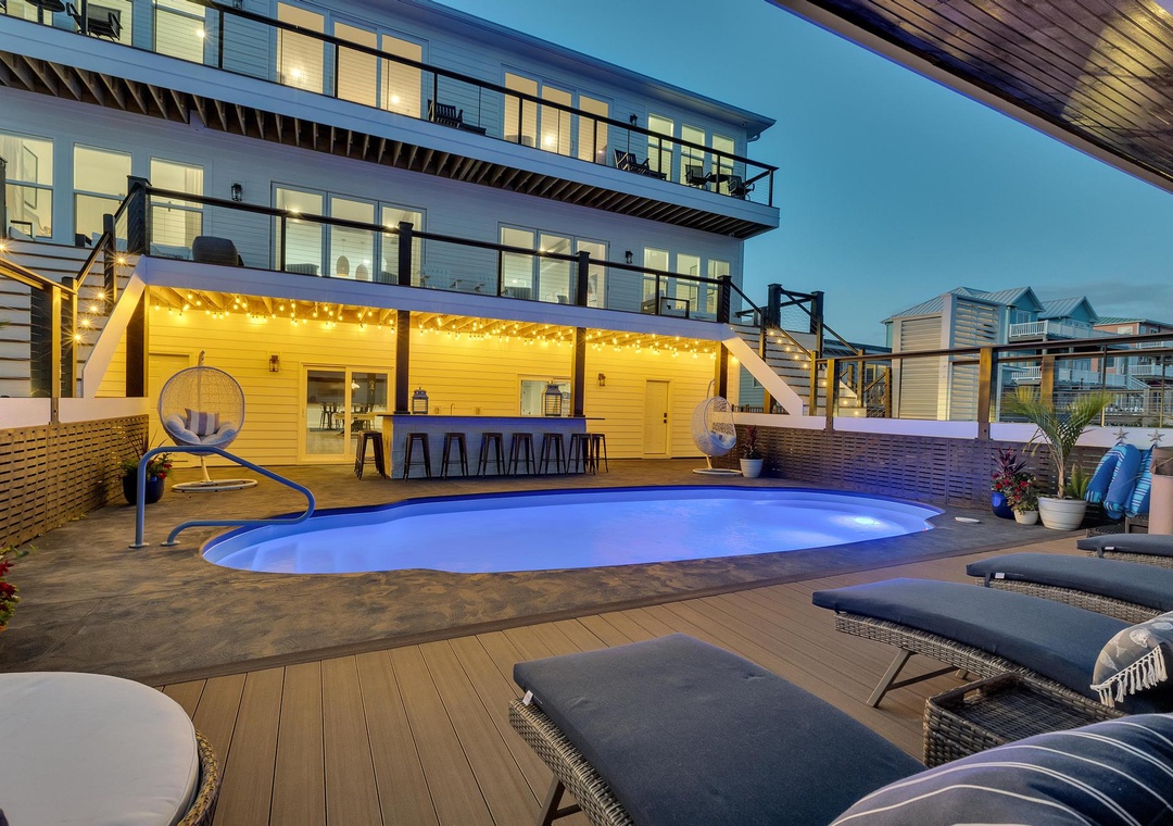 A modern waterfront property featuring an illuminated outdoor entertainment area with pool, lounge seating, and bar access—offering a stylish coastal retreat as evening descends.