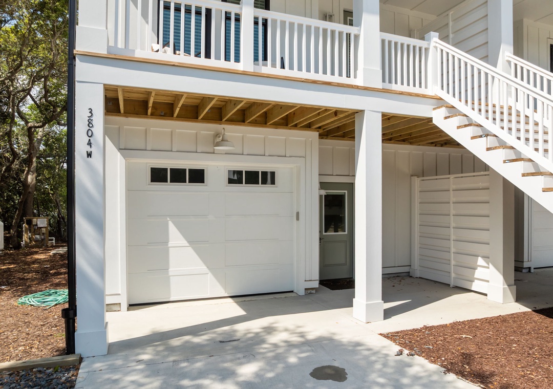 Charming beach house exterior with covered parking and upper deck balcony surrounded by coastal trees.