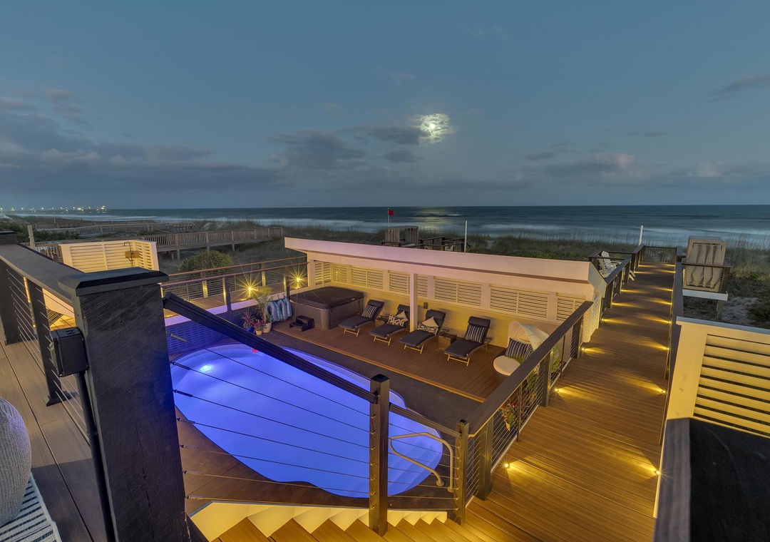 Elevated beachfront deck with illuminated pool, ocean views, and modern outdoor amenities under the evening sky.