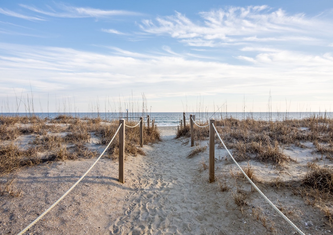 Sandy beach pathway with rope railings leads through dunes to the ocean shoreline.
