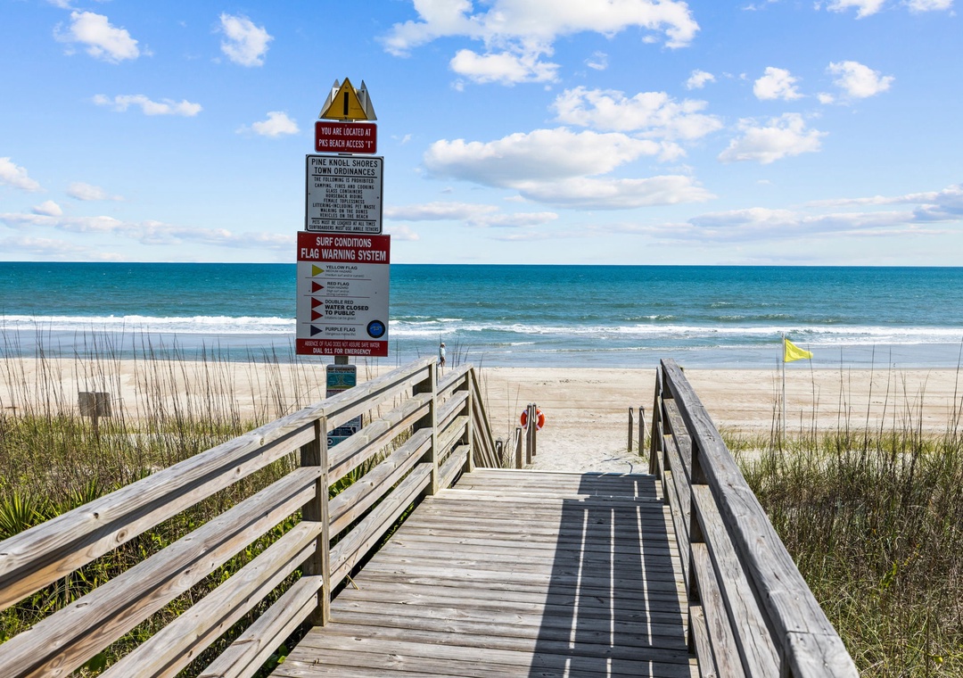 Wooden boardwalk leads to pristine beach with clear blue waters and gentle waves under sunny skies.