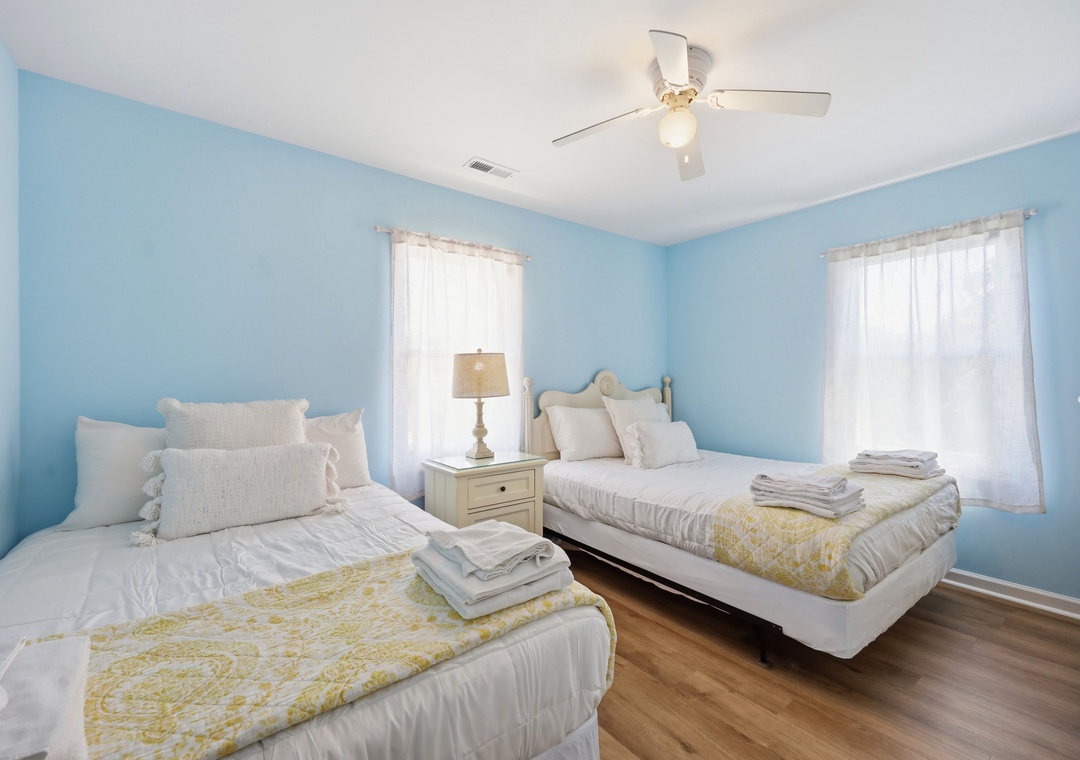 Drift off in this tranquil bedroom with two cozy beds, soft blue walls, natural light, and a gentle ceiling fan overhead keeping you comfortable.