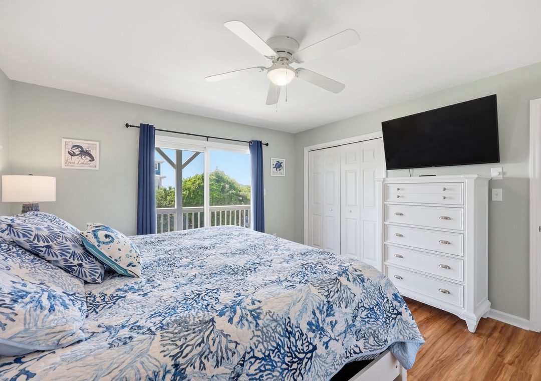 Unwind in your peaceful retreat with a plush bed, ceiling fan, and natural light streaming through windows framing lush greenery. Your flat-screen TV and ample storage await.