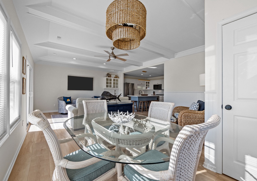 Your bright open-concept living space blends coastal charm with modern comfort, featuring wicker dining chairs and flowing natural light.