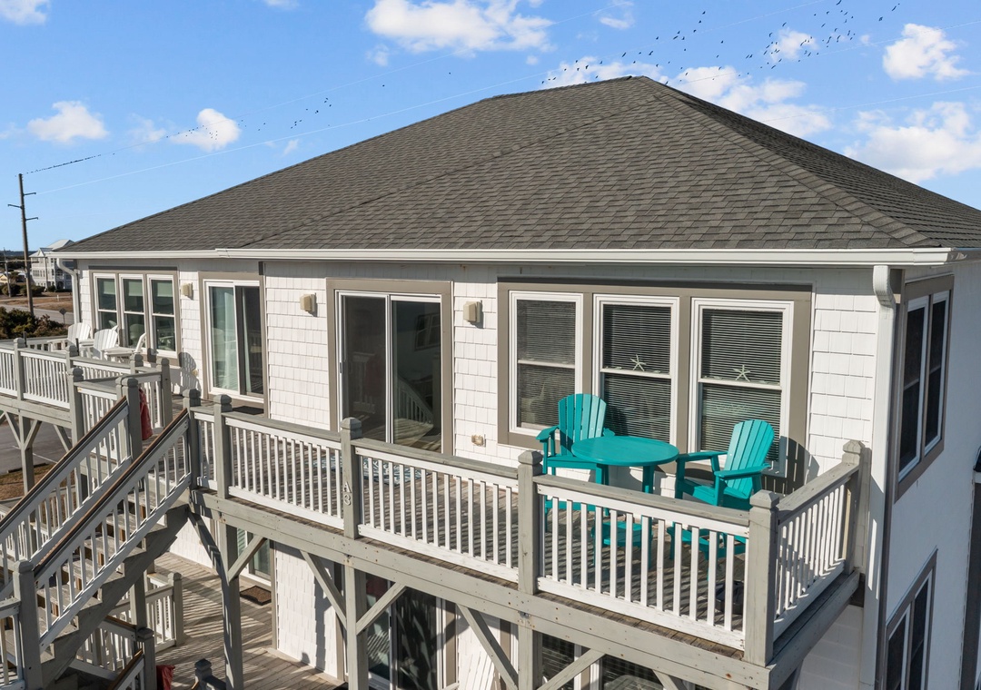Coastal rental property featuring elevated deck with turquoise chairs and white railings under blue skies.