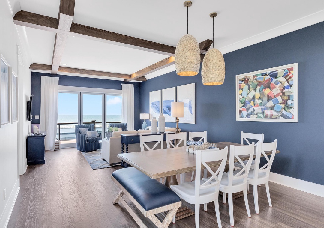 Step into your coastal retreat where rich navy walls meet vaulted ceilings with rustic beams. Your dining space flows seamlessly to a living room framing serene ocean views through floor-to-ceiling windows.
