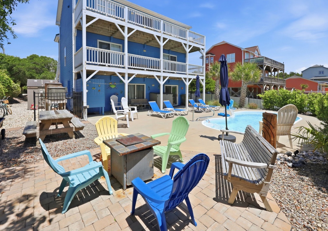 Bright coastal retreat with private pool, sun loungers, colorful Adirondack seating around a fire pit, grilling area, outdoor dining, and shaded lounging—perfect for lazy beach days.