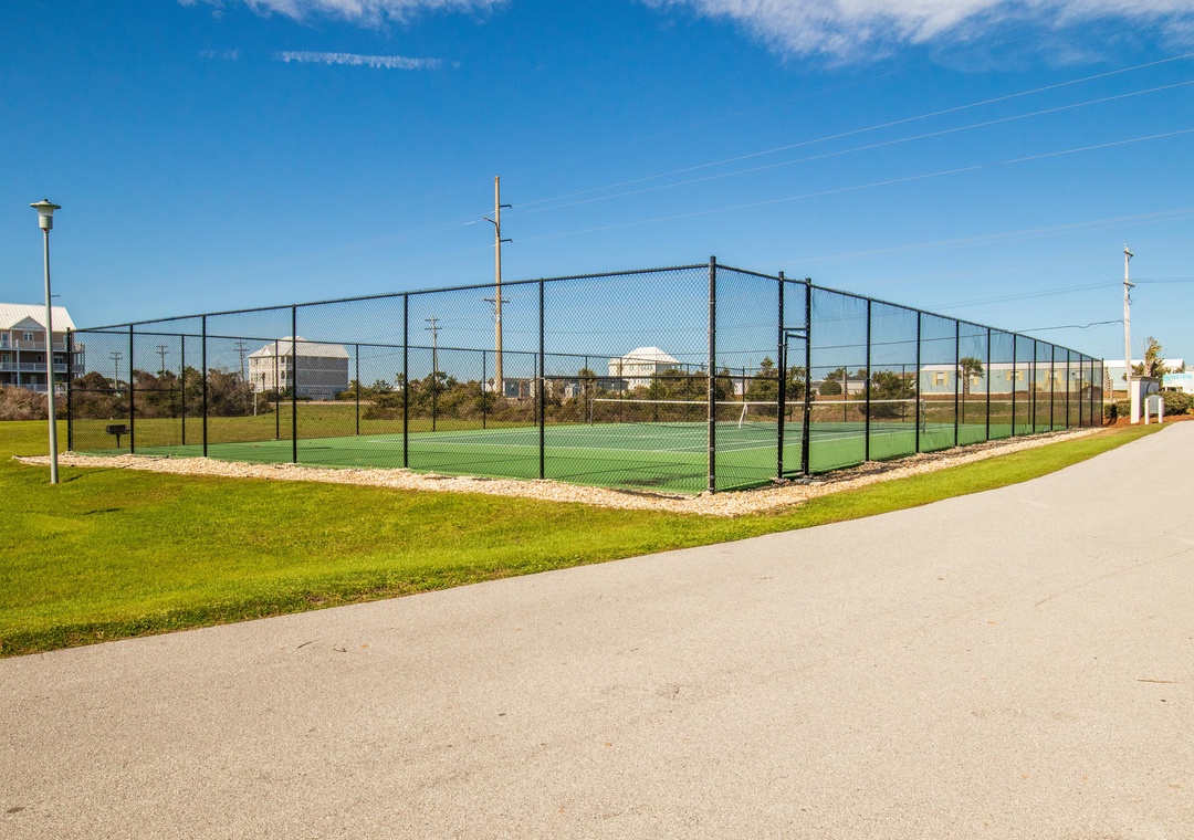 Tennis court with chain-link fencing surrounded by green lawn and residential buildings in the background.