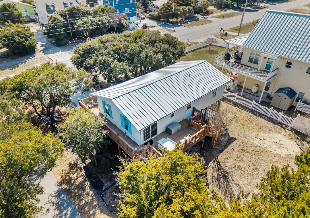 Aerial view of a charming coastal vacation home with metal roof and wraparound deck, nestled among mature trees in a quiet residential neighborhood.