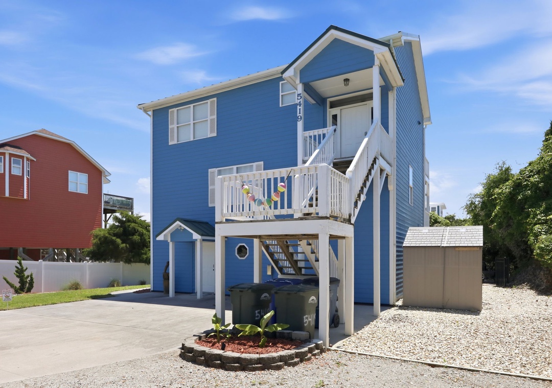 Bright blue coastal beach house with elevated deck and covered parking, located in a quiet residential neighborhood near the shore.