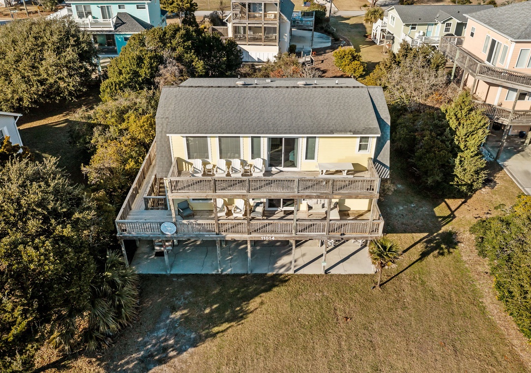 Aerial view of a charming coastal rental property with wraparound decks and outdoor seating, nestled among neighboring beach homes.
