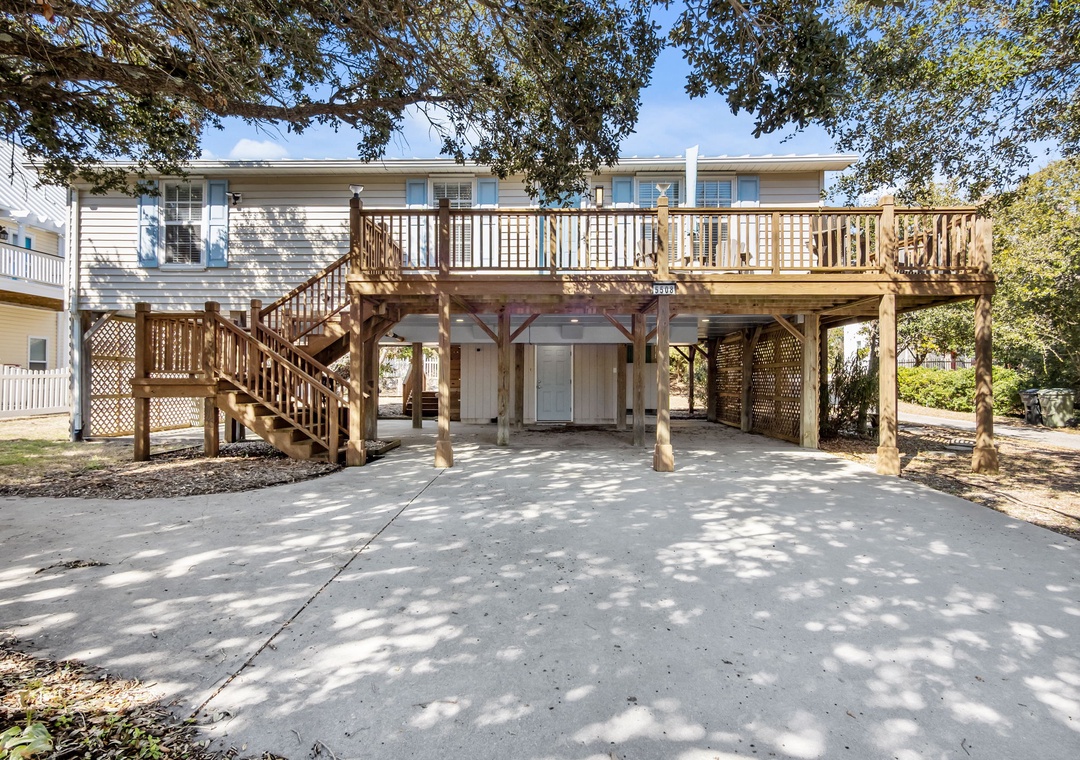 Elevated beach house with spacious deck and covered parking beneath mature trees.