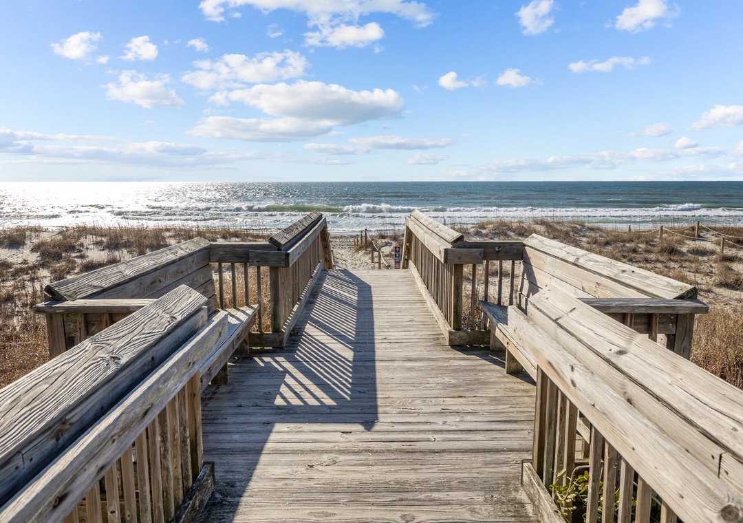Wooden boardwalk leads to pristine sandy beach and rolling ocean waves under a bright blue sky dotted with fluffy white clouds.