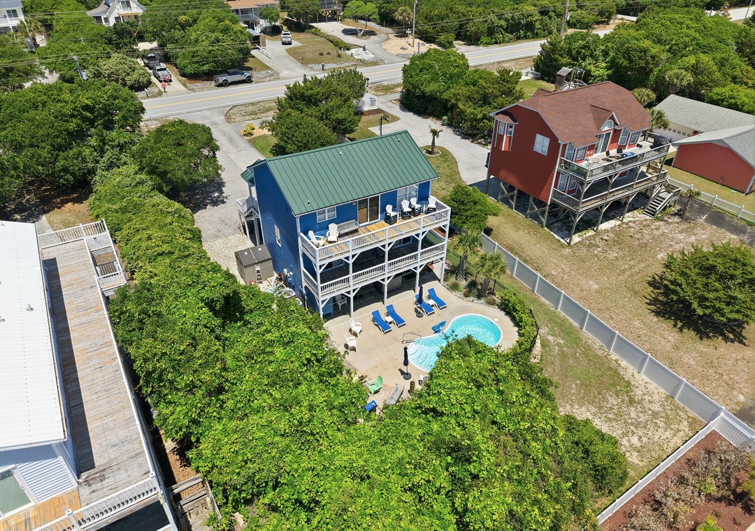 Aerial view of a charming blue coastal vacation rental nestled among lush greenery, featuring a private pool and spacious deck in a peaceful neighborhood setting.