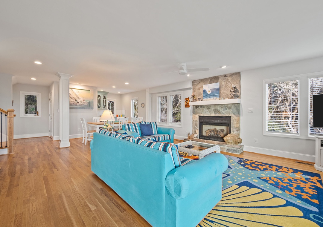 Relax in your bright, coastal-inspired living room with vibrant blue sectional seating, crackling stone fireplace, and panoramic windows overlooking the trees.