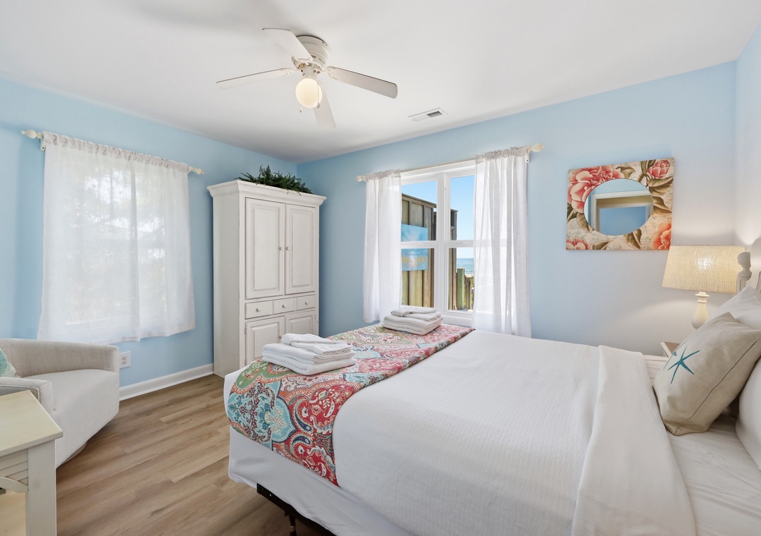 Your serene coastal bedroom features soothing blue walls, crisp white linens, and a ceiling fan to keep you cool. Natural light fills the space, while beach-inspired decor invites relaxation.