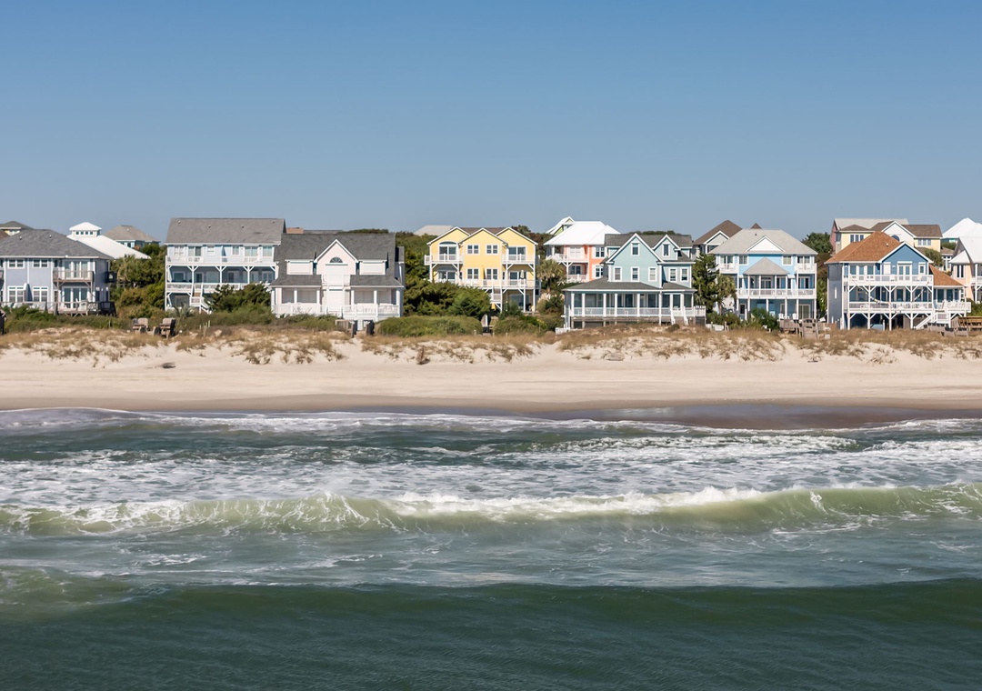 Beachfront vacation homes line a pristine sandy shore, with ocean waves rolling in under clear blue skies. A serene coastal setting perfect for seaside getaways.