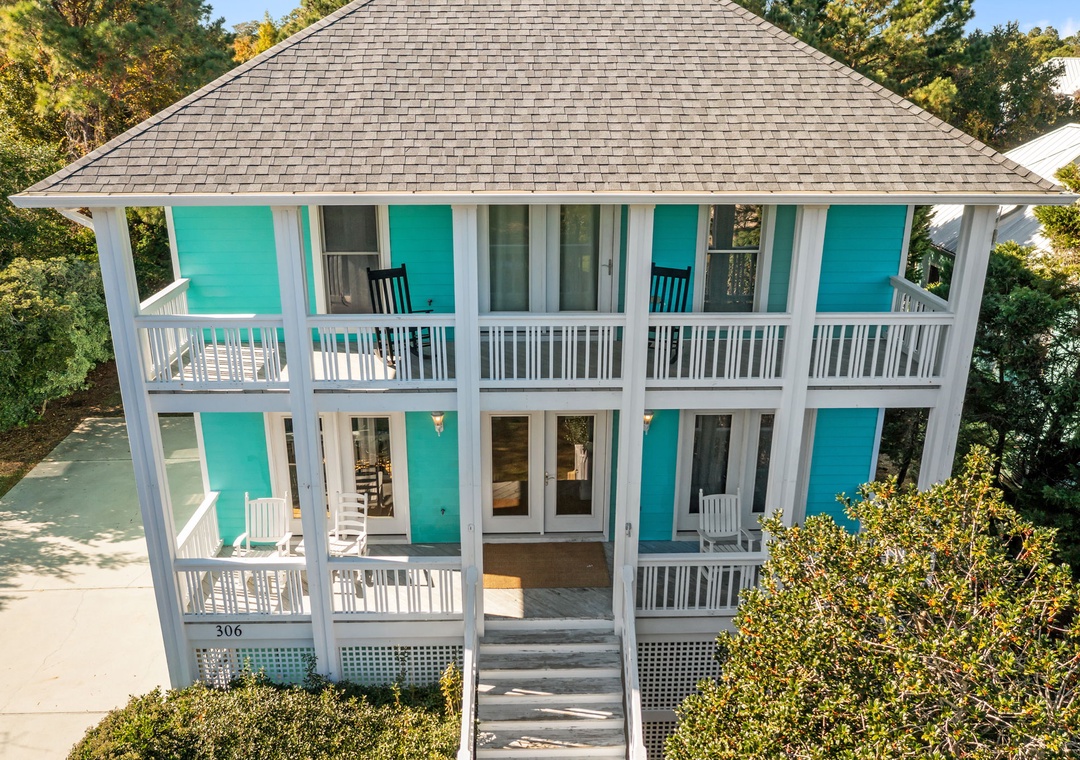 Charming beach cottage with vibrant turquoise shutters and wraparound porches, nestled among lush coastal greenery.