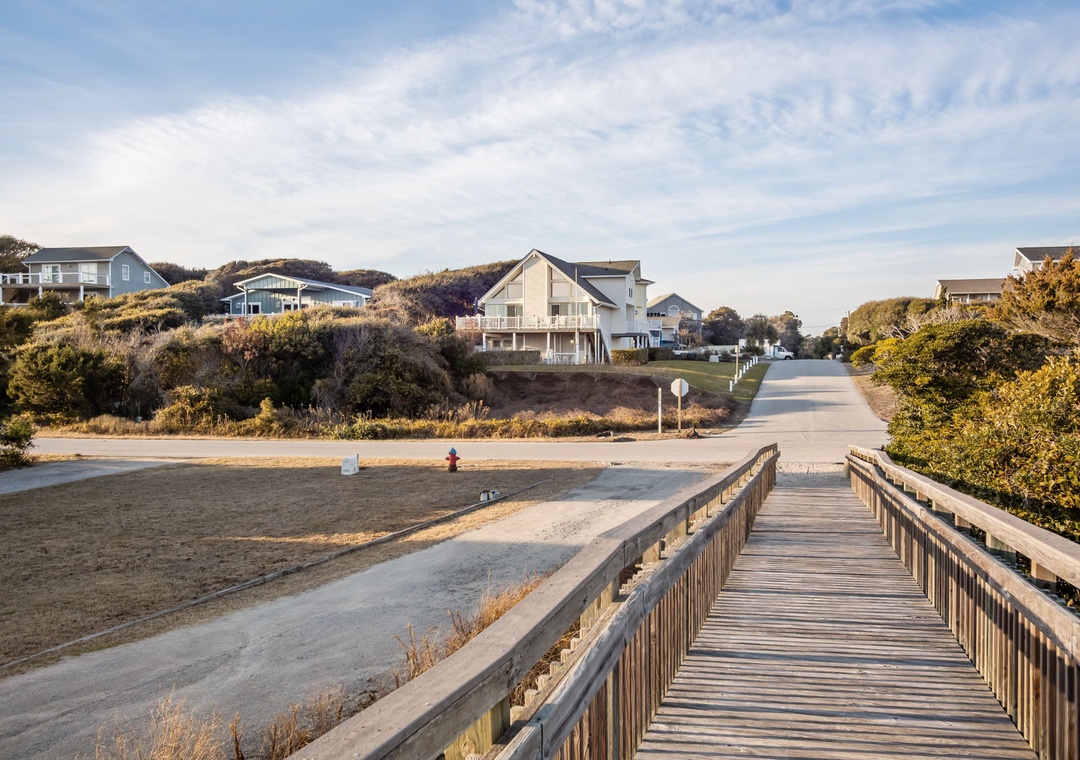 Wooden boardwalk leads through coastal neighborhood with beach houses nestled among dunes and native vegetation.