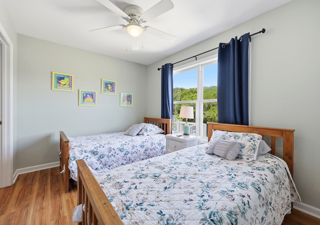 Unwind in your bright, cheerful retreat with two cozy beds dressed in soft florals, a refreshing ceiling fan, and your own balcony access framed by blue curtains.