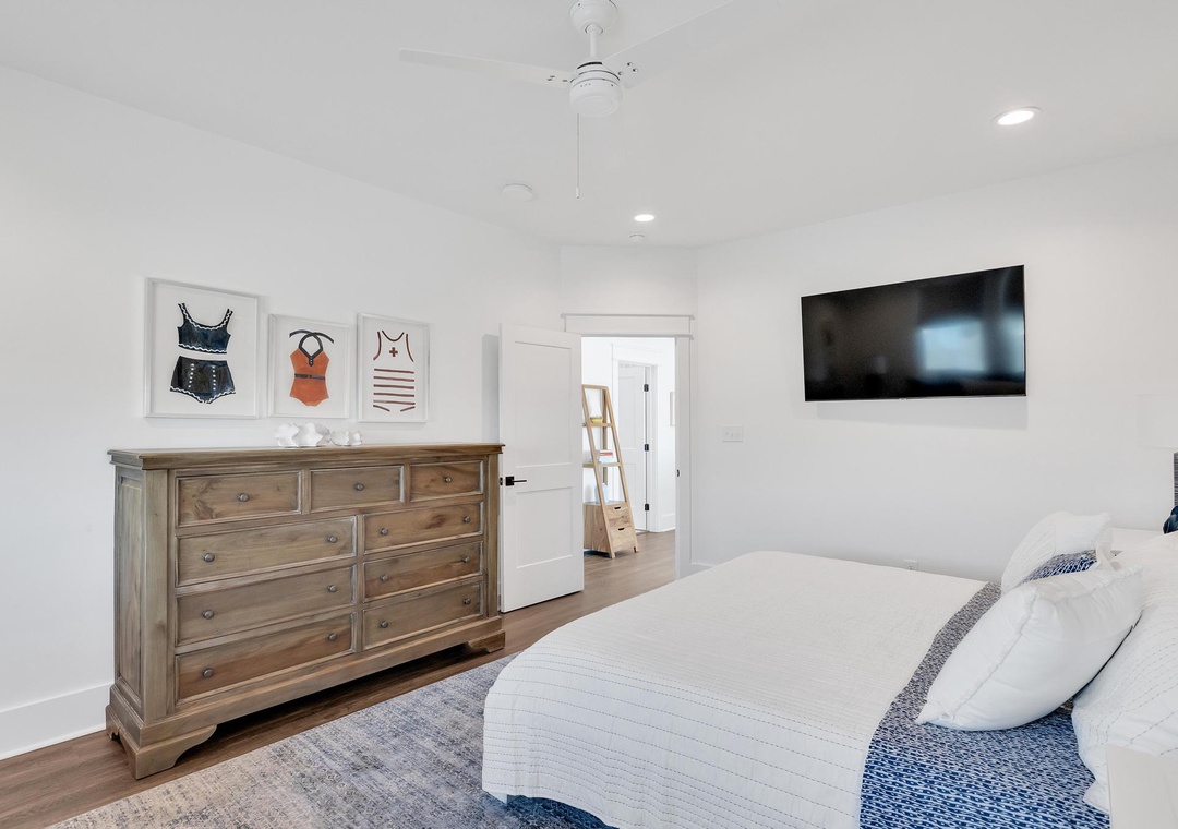 Your bright, serene bedroom awaits with coastal-inspired decor, a plush bed dressed in crisp white linens, and thoughtful touches like a ceiling fan and mounted TV for your comfort.
