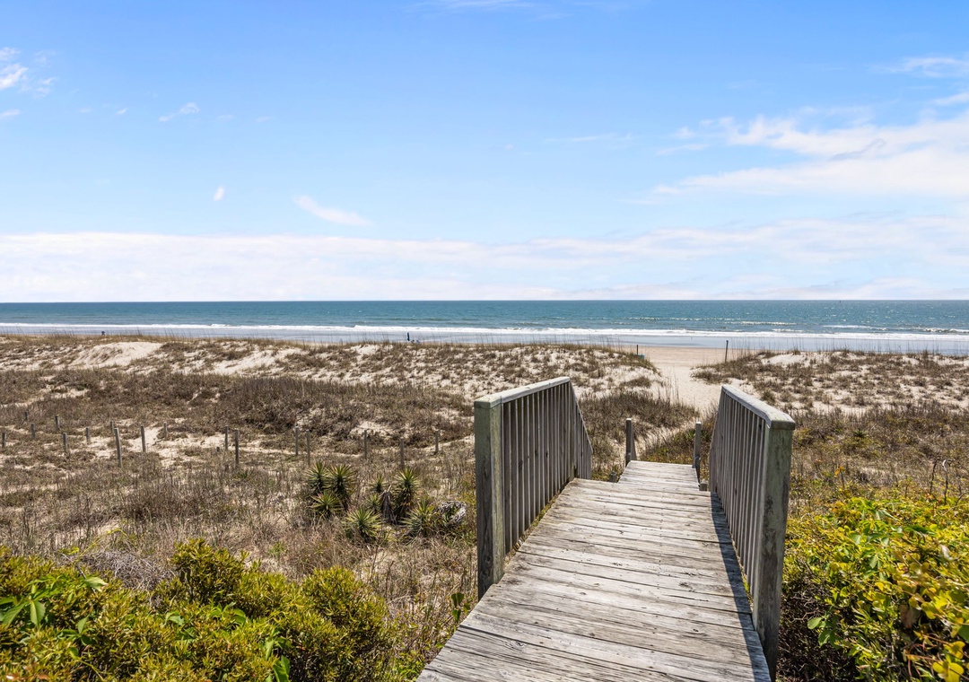 Wooden boardwalk leads through coastal dunes to pristine beach access with ocean views beyond.
