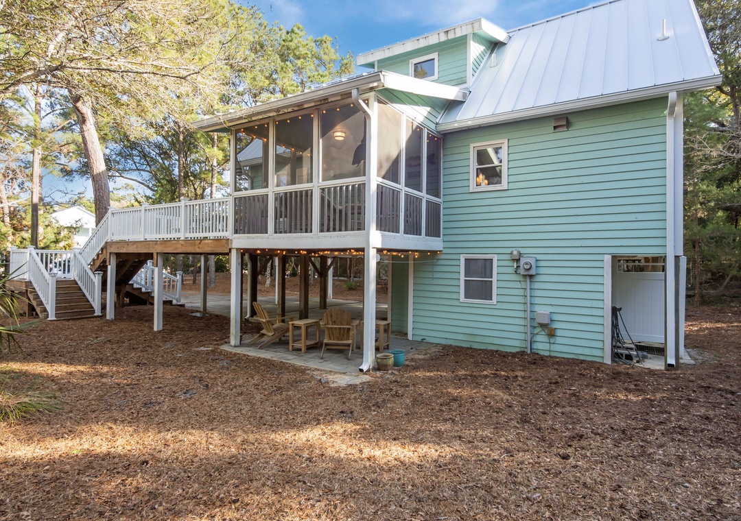 Charming coastal cottage featuring elevated deck with string lights and ground-level patio surrounded by mature trees.