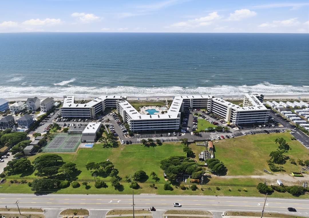 Beachfront property complex featuring multiple mid-rise buildings, swimming pools, tennis courts, and direct ocean access with expansive lawns and parking areas.