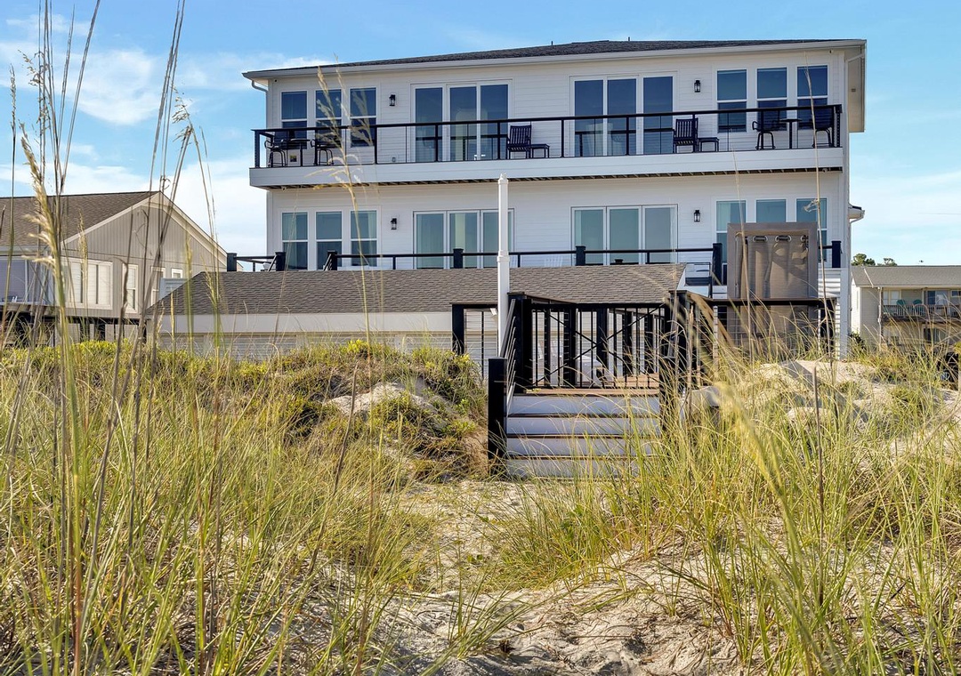 A modern beachfront property with multiple balconies sits just beyond the sandy dunes, offering direct access to coastal relaxation and scenic waterfront surroundings.