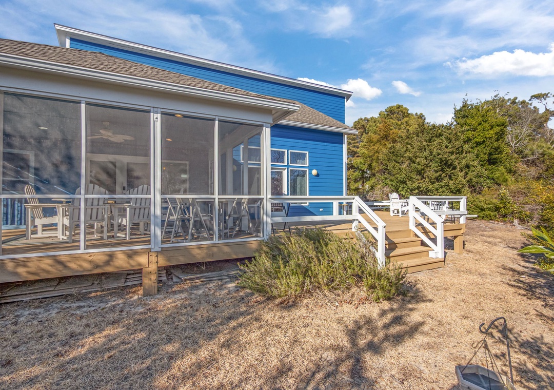 Charming blue coastal cottage with screened porch and deck surrounded by mature trees under bright blue skies.