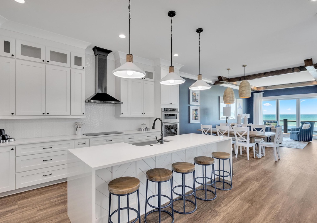 Step into your fully equipped kitchen with gleaming white cabinetry, expansive island seating, and an open layout flowing to the dining area where stunning ocean views await through floor-to-ceiling windows.