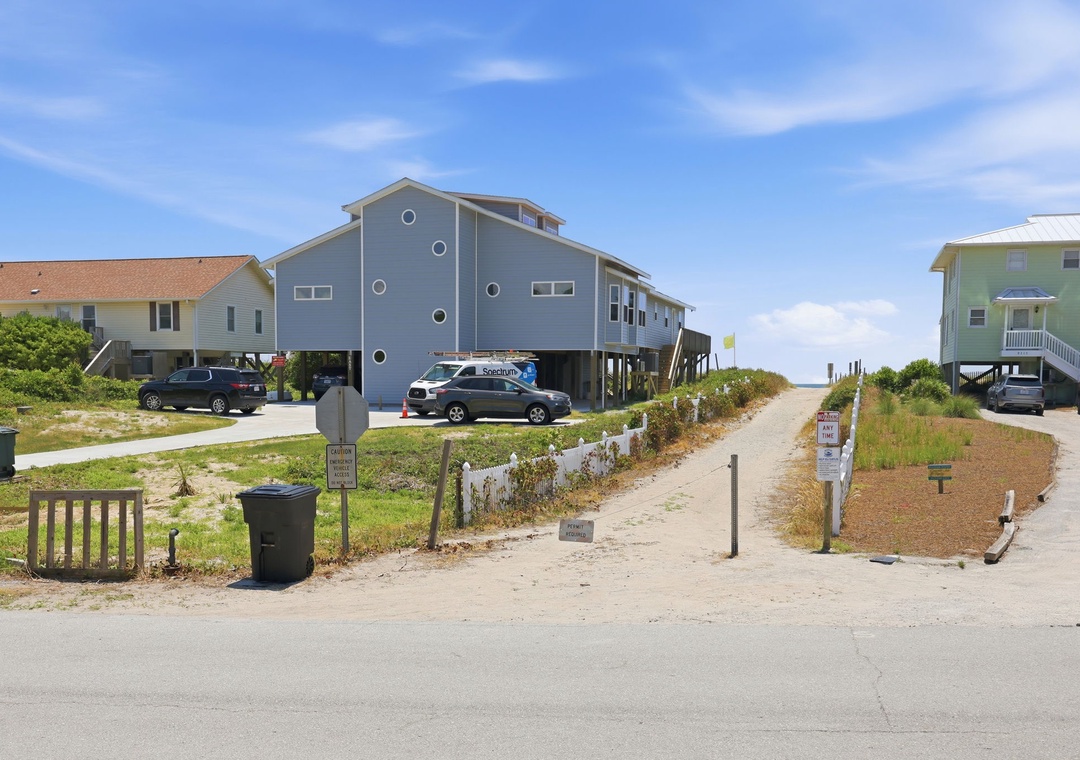 Coastal vacation homes elevated on stilts with direct beach access through a sandy pathway, surrounded by natural dune vegetation under clear blue skies.