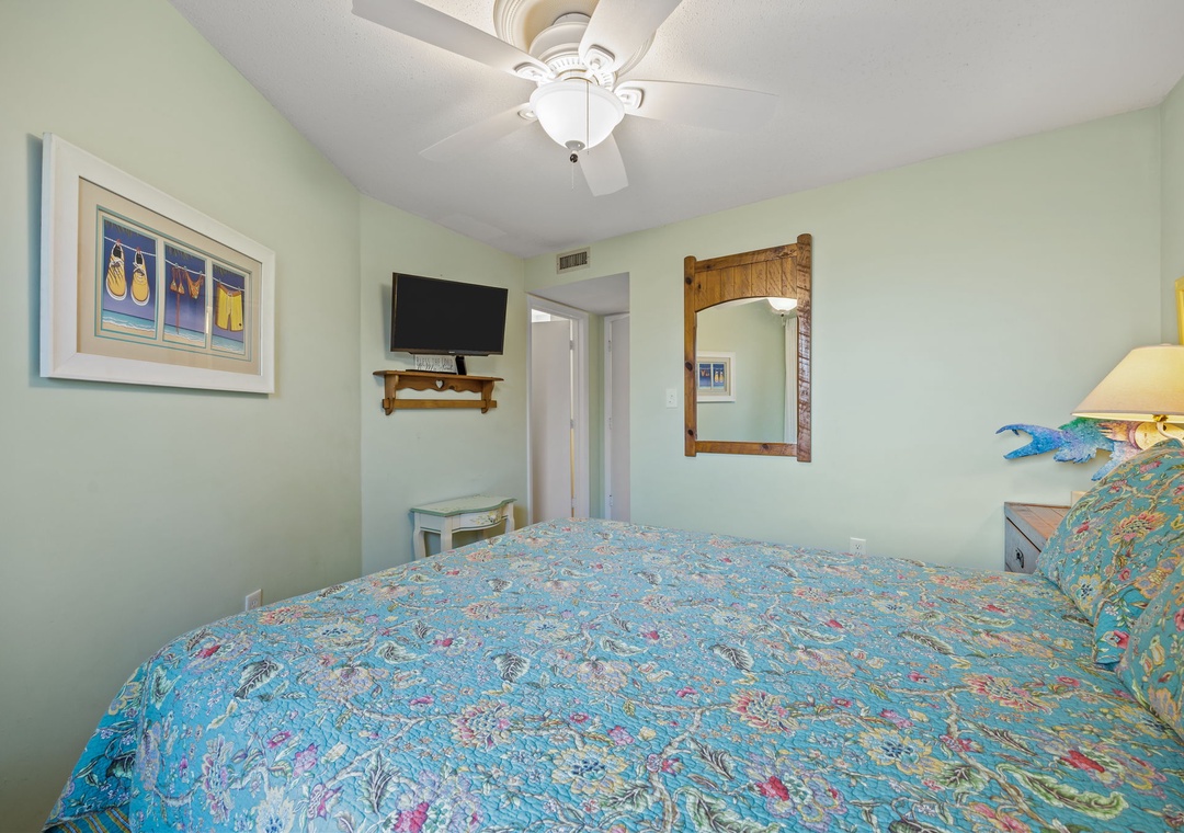 Unwind in your peaceful bedroom featuring tropical-themed linens, a ceiling fan for comfort, and entertainment at your fingertips with the mounted TV.