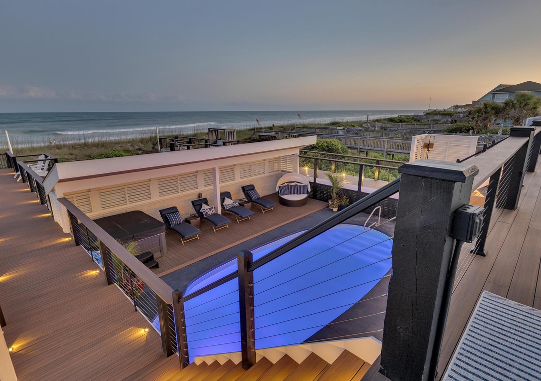 Oceanfront property with expansive deck featuring an illuminated pool, lounge seating, and direct beach access via wooden walkway.