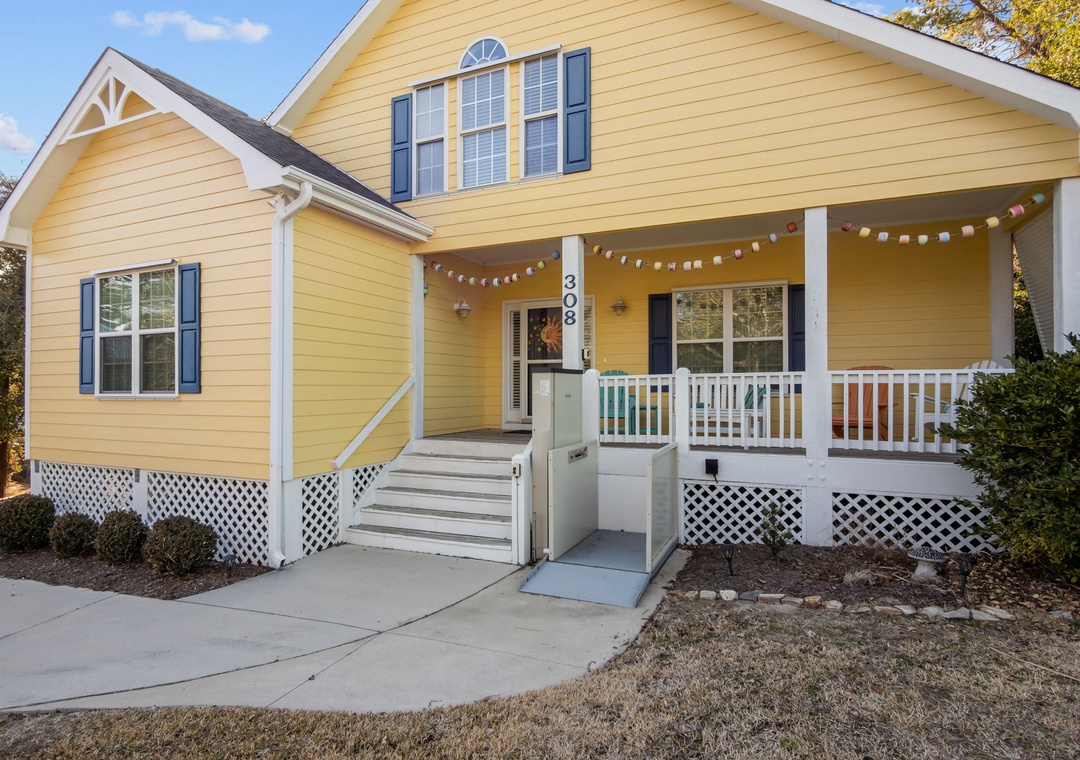 Charming yellow cottage with welcoming front porch and accessibility ramp, featuring festive string lights and comfortable outdoor seating area.