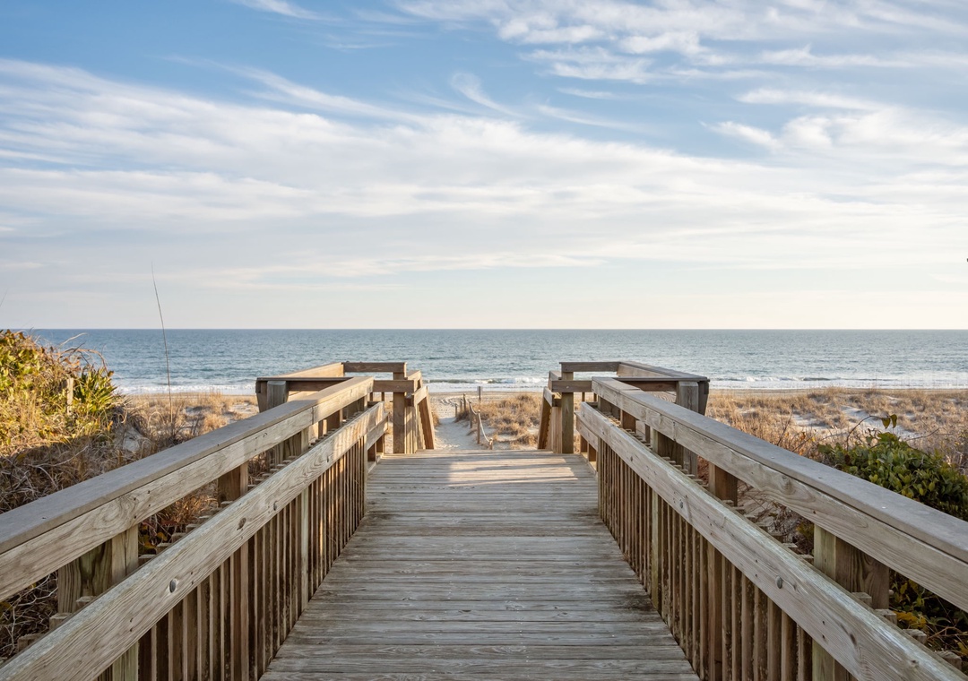 Wooden boardwalk leads to pristine beach with ocean waves and coastal dunes under expansive sky.